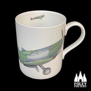 May include: White ceramic mug with a green and blue camouflage design of a vintage airplane. The mug has a handle and the words "FOLEY POTTERY" are printed on the bottom.