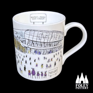 Könnte beinhalten: Weiße Keramiktasse mit einer Schwarzweißillustration eines Stadions mit dem Text "Bramley-Moore Dock Stadium" an der Seite. Das Stadion ist von kleinen Strichmännchen in verschiedenen Farben umgeben.