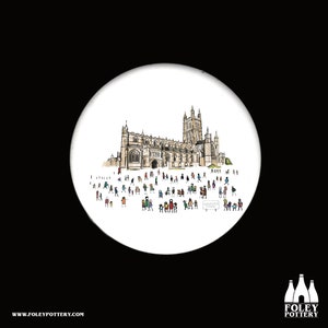 Puede incluir: Un posavasos de cerámica blanca con una ilustración dibujada a mano de una catedral con una multitud de personas delante. El texto "GLOUCESTER CATHEDRAL" está impreso en un cartel en primer plano.