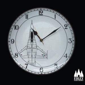 Könnte beinhalten: Weiße Keramikuhr mit schwarzen Zeigern und schwarzem Rand. Das Zifferblatt der Uhr zeigt eine Schwarzweißillustration eines Kampfjets. Die Zahlen 1 bis 12 sind in Schwarz um das Zifferblatt gedruckt.
