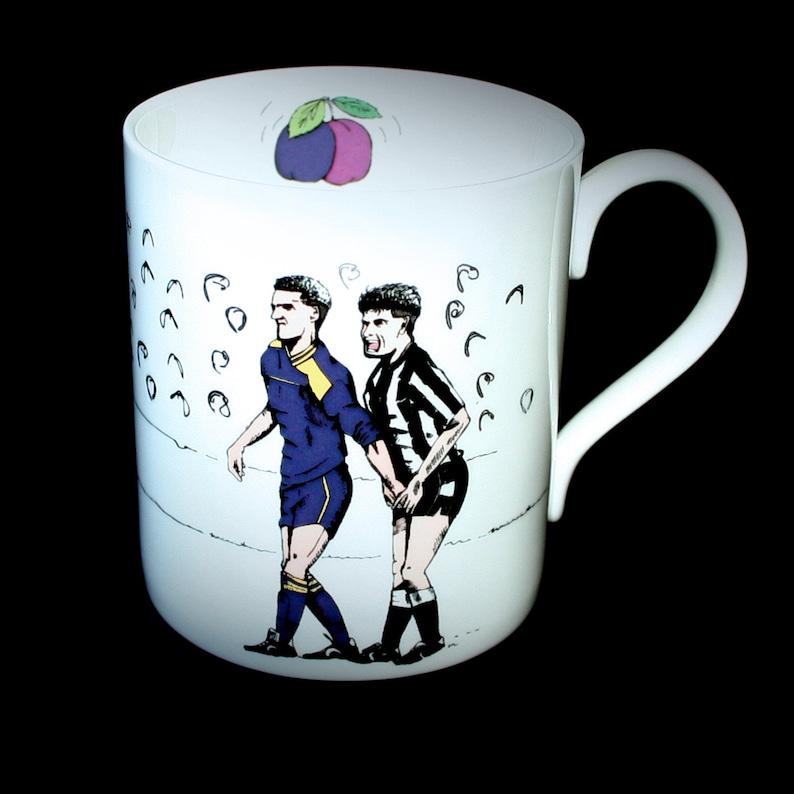 May include: White ceramic mug with a cartoon illustration of two soccer players, one in a purple and yellow uniform and the other in a black and white striped uniform. The mug has a plum design on the top.