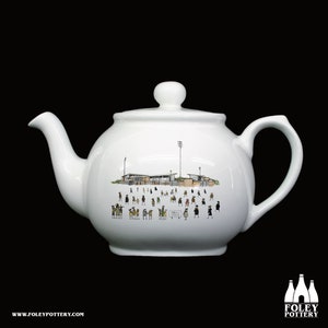 Könnte beinhalten: Weiße Keramik-Teekanne mit einer detaillierten Illustration einer Stadionszene, die eine Menschenmenge und Stadionlichter zeigt. Die Teekanne hat einen abgerundeten Körper, einen gebogenen Griff und einen Ausguss. Auf der Unterseite der Teekanne steht "www.foleypottery.com".