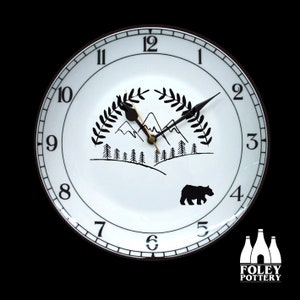 May include: White ceramic clock with black numbers and hands. The clock face features a black and white illustration of mountains, trees, and a bear. The clock is framed with a black rim and has a gold centre.