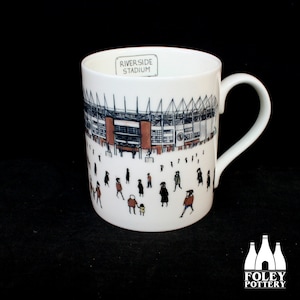 May include: White ceramic mug with a black and white illustration of the Riverside Stadium, home of Middlesbrough FC. The stadium is depicted with a crowd of people in front of it. The mug has the words "Riverside Stadium" printed on the side.