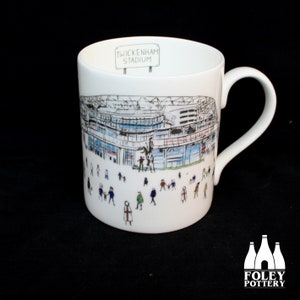 Puede incluir: Taza de cerámica blanca con una ilustración en blanco y negro del estadio de Twickenham. El estadio está rodeado de figuras de palitos de personas. La taza tiene un asa negra y las palabras "Twickenham Stadium" están impresas en la parte superior de la taza.