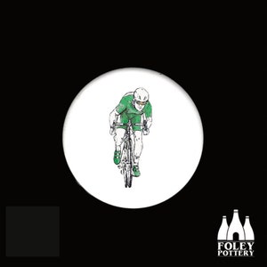 May include: A white ceramic button featuring a black and white line drawing of a cyclist wearing a green jersey and a white helmet riding a bicycle.