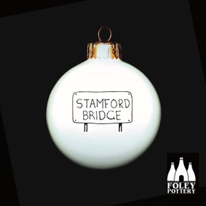May include: White Christmas ornament with a black and white sign that reads "Stamford Bridge". The ornament is made by Foley Pottery.