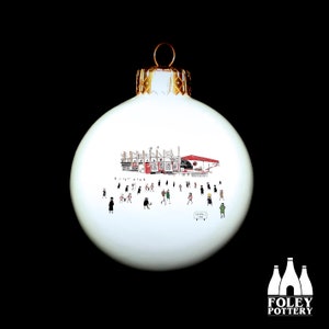 May include: White ceramic Christmas ornament with a hand-drawn illustration of a stadium with people in front of it. The ornament has a gold top and a red and white stadium.