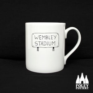 May include: White ceramic mug with a black and white illustration of a sign that reads "Wembley Stadium".