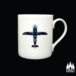 Könnte beinhalten: Eine weiße Keramiktasse mit einem Flugzeugdesign in Schwarz und Blau. Das Flugzeug ist nach unten gerichtet, die Flügel sind ausgebreitet.