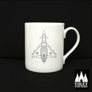 Könnte beinhalten: Weiße Keramiktasse mit einer schwarzen Linienzeichnung eines Eurofighter Typhoon Jets auf der Vorderseite. Der Jet ist nach oben gerichtet, mit ausgebreiteten Flügeln. Die Tasse hat einen schwarzen Henkel.