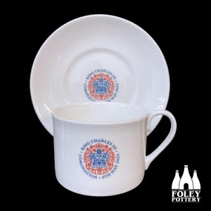 May include: White teacup and saucer with a blue and red design commemorating the coronation of King Charles III on May 6, 2023. The design features a crown and floral motifs.