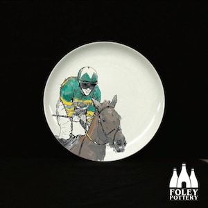 May include: A white ceramic plate featuring a black and white illustration of a jockey riding a horse. The jockey is wearing a green and yellow racing suit and a helmet. The horse is brown and white.