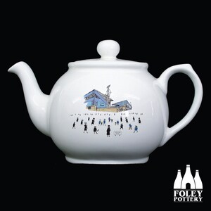 May include: White ceramic teapot with a blue and white illustration of a stadium and people walking around it. The text "GOODISON PARK" is visible on the teapot.