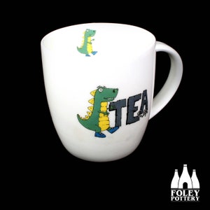 Puede incluir: Taza de cerámica blanca con un dinosaurio verde sosteniendo la palabra "TEA" en letras grises. El dinosaurio lleva zapatos azules y tiene detalles amarillos. Hay un dinosaurio más pequeño en el fondo.