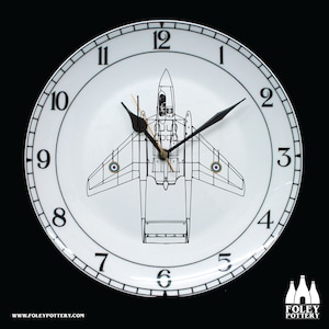 May include: A white ceramic clock with black numbers and hands. The clock face features a black and white line drawing of a jet aircraft. The clock is 12 inches in diameter.