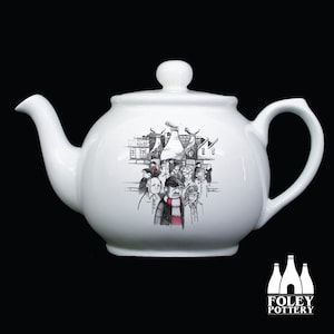 May include: White ceramic teapot with a black and white illustration of a group of people standing in front of a building. The illustration includes a red and white scarf. The teapot is made by Foley Pottery.