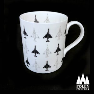 May include: White ceramic mug with a black and white pattern of fighter jets. The mug has a handle and the text "FOLEY POTTERY" is printed on the bottom.
