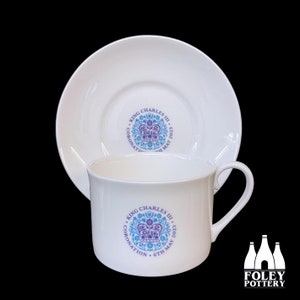 May include: White teacup and saucer with a blue and purple design commemorating King Charles III's coronation on May 6, 2023. The design features a crown and floral motifs. The text "KING CHARLES III CORONATION 6TH MAY 2023" is printed on the cup and saucer.