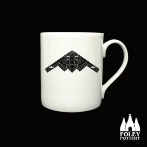 May include: A white ceramic mug with a black silhouette of a B-2 Spirit stealth bomber aircraft. The mug has a handle on the right side.