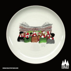 May include: White ceramic plate with a cartoon illustration of a crowd of people wearing red and white scarves watching a soccer game at the Emirates Stadium.