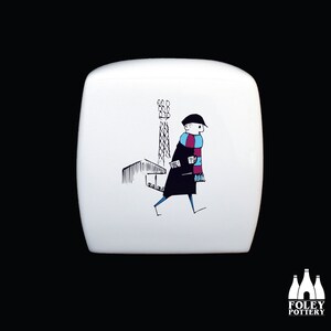 May include: White ceramic mug with a black and white illustration of a person wearing a blue and pink scarf walking past a stadium floodlight.