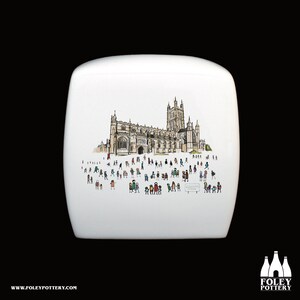Puede incluir: Tapa de tanque de inodoro de cerámica blanca con una ilustración en blanco y negro de una catedral y personas frente a ella. El texto "GLOUCESTER CATHEDRAL" está escrito en un cartel que sostiene una de las personas de la ilustración.