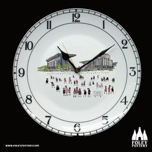 May include: A white ceramic clock plate with black numbers and hands. The clock face features a black and white illustration of a stadium with people in pink and black clothing. The text "CHASE STADIUM" is visible on the clock face.