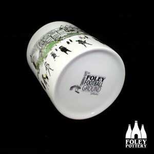 Peut inclure: Une tasse en c&eacute;ramique blanche avec une illustration en noir et blanc d'un stade de football et de personnes sur le c&ocirc;t&eacute;. Le fond de la tasse porte le texte "The Foley Football Ground Collection" en noir.