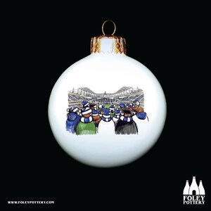 Puede incluir: Adorno navideño blanco con una ilustración dibujada a mano de una multitud de estadio con gorros y bufandas azules y blancas. La multitud está de espaldas al espectador, mirando un partido de fútbol.