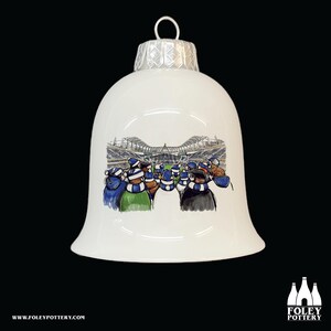 Puede incluir: Adorno navideño de cerámica blanca con forma de campana con una ilustración azul y blanca de una multitud de estadio. El adorno tiene una parte superior blanca y plateada.