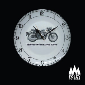 May include: A white ceramic clock with black numbers and a black hand. The clock face features a black and white illustration of a Velocette Venom motorcycle with the text "Velocette Venom 1955 500cc".