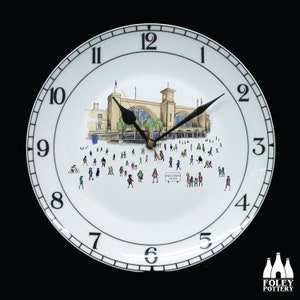 Puede incluir: Un reloj de cerámica blanca con números y manecillas negros. La esfera del reloj presenta una ilustración en blanco y negro de la estación de King's Cross en Londres, Inglaterra. La ilustración incluye personas caminando alrededor de la estación.