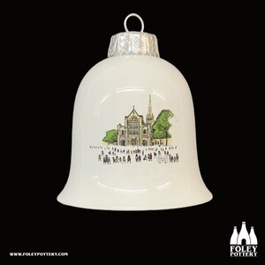 May include: White ceramic Christmas ornament shaped like a bell with a silver top. The ornament features a hand-drawn illustration of a church with people in front of it. The text "St. Mary's Church, Saffron Walden" is visible on the sign.