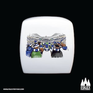 May include: White ceramic mug with a cartoon illustration of a crowd of people wearing blue and white scarves, looking towards a stadium in the background. The stadium is a large, modern structure with a white roof and a green field.