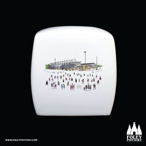 Peut inclure: Couverture de réservoir de toilette en céramique blanche avec une illustration en noir et blanc d'un stade avec des personnes au premier plan. Le stade a un toit blanc et un terrain vert. Les personnes portent des vêtements de différentes couleurs.