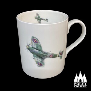 May include: White ceramic mug with a green and brown airplane illustration. The airplane has red, white, and blue markings. The mug has a handle on the right side.