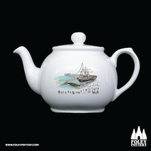 Puede incluir: Tetera de cerámica blanca con una ilustración pintada a mano de un barco atracado en un muelle con gente caminando por el paseo marítimo.