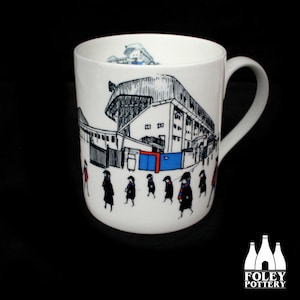 以下が含まれることがあります： 白地に黒と白のイラストが描かれたセラミック製のマグカップ。スタジアムと、スタジアムに向かって歩く人々が描かれている。スタジアムは、青と赤のゲートのあるシンプルなスタイルで描かれている。マグカップには黒い取っ手がある。