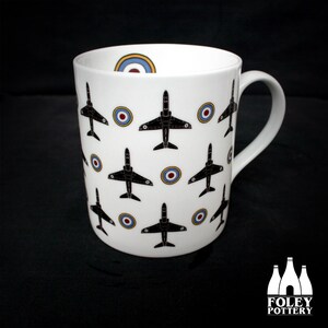 May include: White ceramic mug with a black and white repeating pattern of jet planes and red, white, and blue roundels. The mug has a black handle.