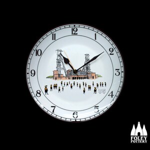 May include: A white ceramic clock with black hands and numbers. The clock face features a black and white illustration of a coal mine with the text "Clipstone Colliery".