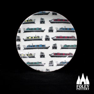 Peut inclure: Une assiette en céramique blanche avec un motif répétitif de bateaux de canal colorés. Les bateaux sont noirs avec des toits et des fenêtres de différentes couleurs. L'assiette est ronde et a un bord légèrement surélevé.