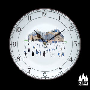 Puede incluir: Un reloj de cerámica blanca con un borde negro y marrón. La esfera del reloj presenta una ilustración en blanco y negro de un estadio con gente caminando alrededor. Los números en la esfera del reloj son negros y las manecillas son negras. El reloj está hecho por Foley Pottery.