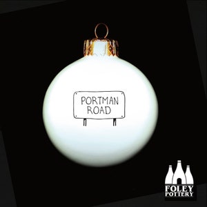 May include: White Christmas ornament with black hand-drawn lettering that reads "Portman Road". The ornament has a gold top and is on a black background.