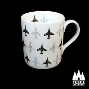 May include: A white ceramic mug with a black and white pattern of fighter jets. The mug has a handle and the text "FOLEY POTTERY" is printed on the bottom.