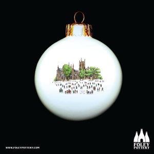 以下が含まれることがあります： 金色のキャップが付いた白いクリスマスオーナメント。人々や木々のある大聖堂の詳細なイラストが描かれています。オーナメントは光沢のある仕上がりで、黒い背景に配置されています。