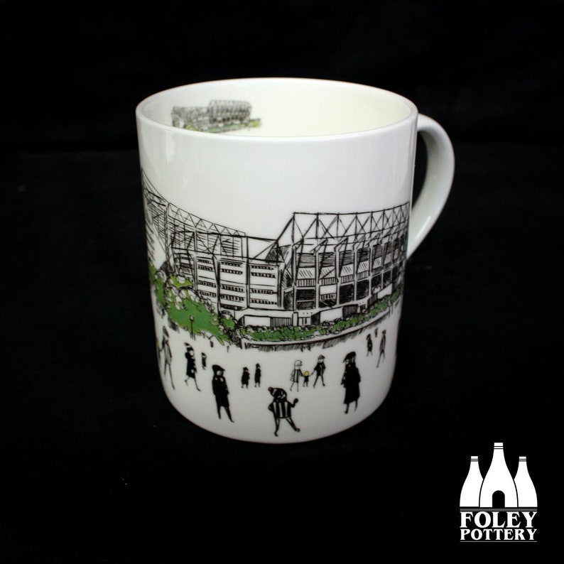 Peut inclure: Une tasse en c&eacute;ramique blanche avec une illustration en noir et blanc d'un stade et de personnes qui marchent. La tasse a une poign&eacute;e noire et les mots "FOLEY POTTERY" sont imprim&eacute;s au fond.