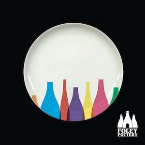 Pode incluir: Um prato de cerâmica branco com um design colorido de garrafas em fila ao longo da borda inferior. As garrafas são em tons de marrom, azul, vermelho, roxo, amarelo, rosa e verde. O prato faz parte de um conjunto de louça da Foley Pottery.