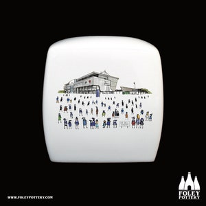 May include: White ceramic toilet tank lid featuring a hand-drawn illustration of a stadium with a crowd of people in front of it. The stadium is light grey with a red roof. The people are drawn in a simple style wearing blue and black clothing. The text "AZ Bell Stadium" is visible on a sign in the image.