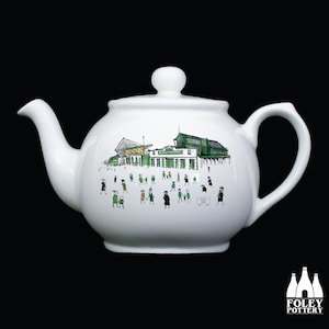 Puede incluir: Tetera de cerámica blanca con tapa y asa. La tetera presenta una ilustración dibujada a mano de un estadio con gente vestida de verde y negro. Las palabras "HOPE PARK" están en un letrero. El logotipo de Foley Pottery está en la parte inferior.